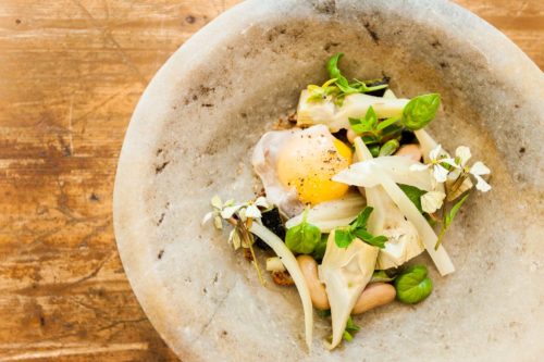 A stone bowl containing cooked artichoke hearts, a duck egg, beans, edible flowers and herbs on a wooden table top, served at The Dairy restaurant