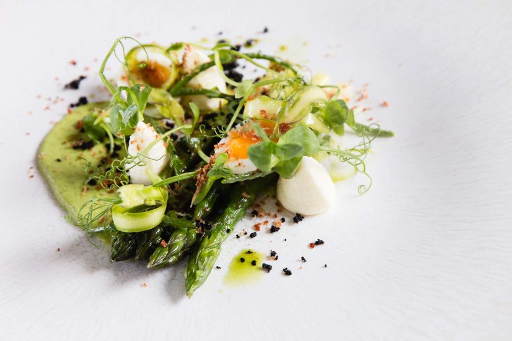 A fine dining plate of shaved and whole asparagus with an egg, pea sprouts and a pea foam, on a white textured plate