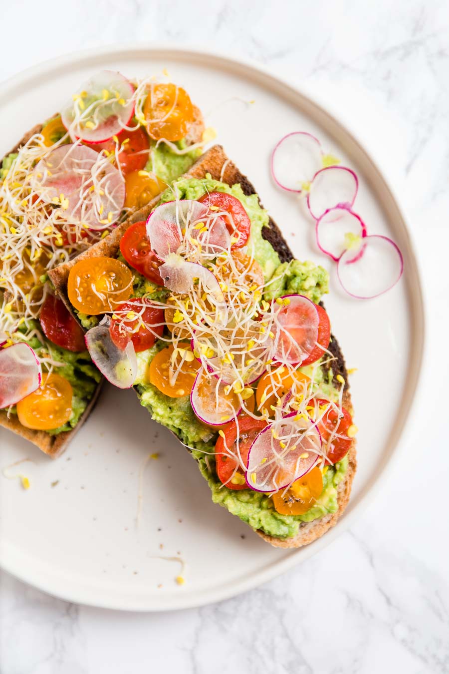 Slices of artisan bread toasted, spread with avocado, topped with sliced cherry tomatoes, radish and bean sprouts on a white, shallow plate