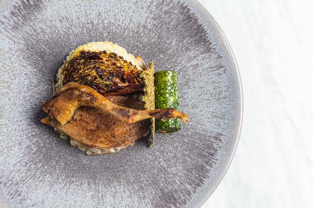 Chicken leg carefully plated in a fine dining style, with a leaf wrapped confit leg meat on a textured grey plate