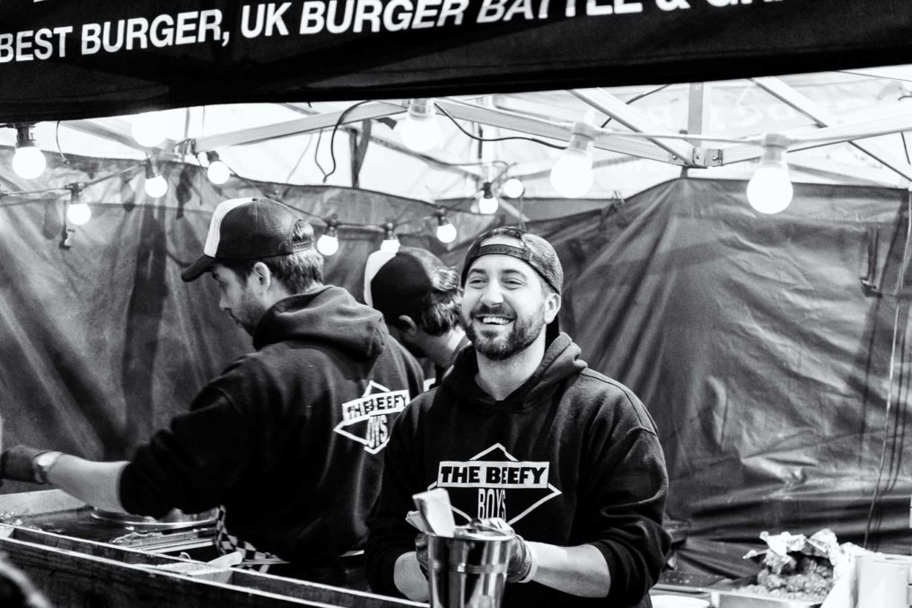 Beefy Boys serving street food. Bearded male smiles big at customers who are out of shot