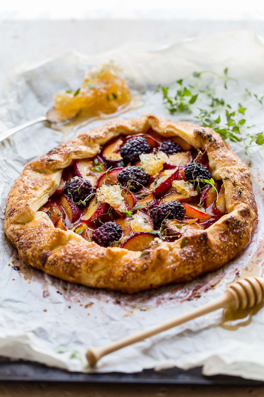 Round cornmeal pastry galette filled with roasted plums and blackberries, drizzled with honey, sitting on a greaseproof sheet with green micro herbs and honeycomb