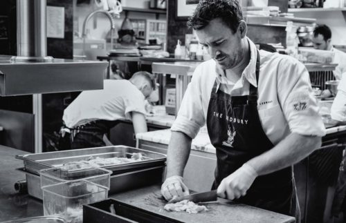 Black and white photo of chef Greg Austin slicing meat in The Ledbury kitchen with kitchen activity in the background