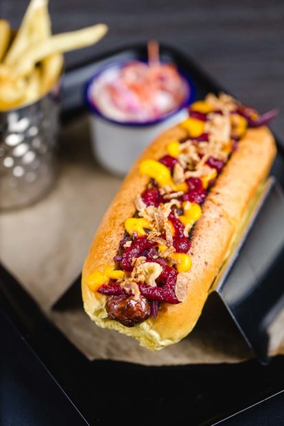 A chilli hotdog dressed with a spicy tomato ketchup, mustard and crispy onion pieces with a container of fries and tub of coleslaw in the background. Served at The Bootroom at LFC Anfield