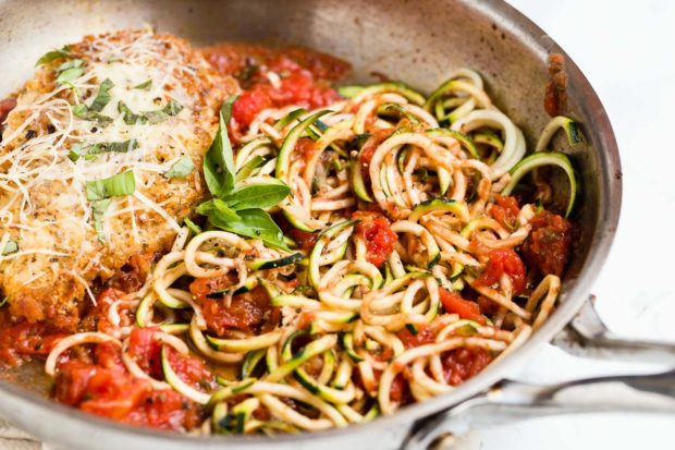 A frying pan with a cooked crispy chicken breast covered with herbs and parmesan cheese next to cooked tomatoes and zucchini noodles or zoodles