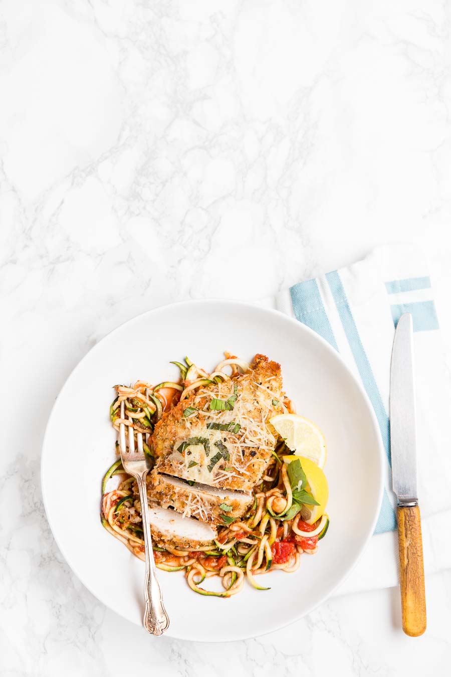 Baked chicken breast on a bed of zucchini noodles or zoodles with herbs and parmesan cheese on a white plate and a blue striped cloth with rustic knife and fork