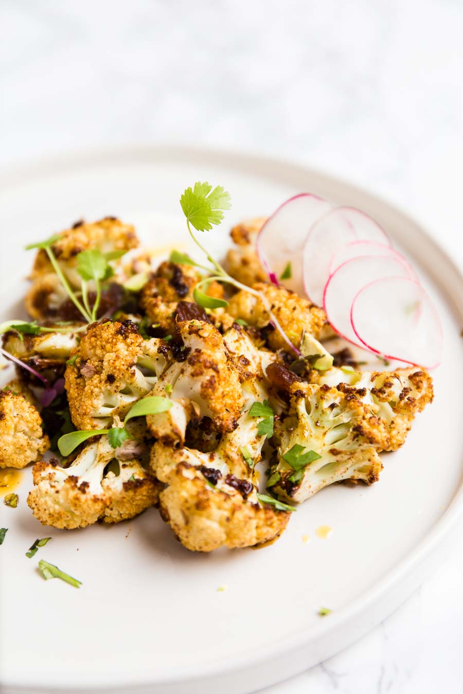 Cooked curried cauliflower served on a shallow white plate, garnished with herbs and wafer thin slices of radish