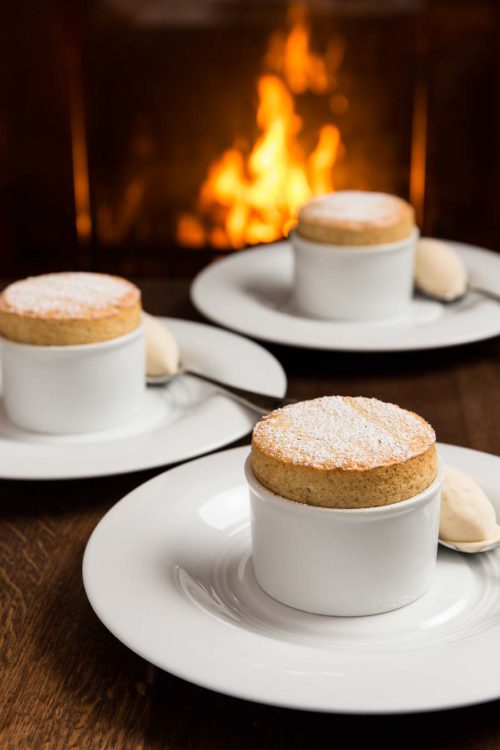 Three soufflé in white bowl, on white plates, along side spoons with quenelle ice-cream, on a wooden table with a lit fireplace in the background