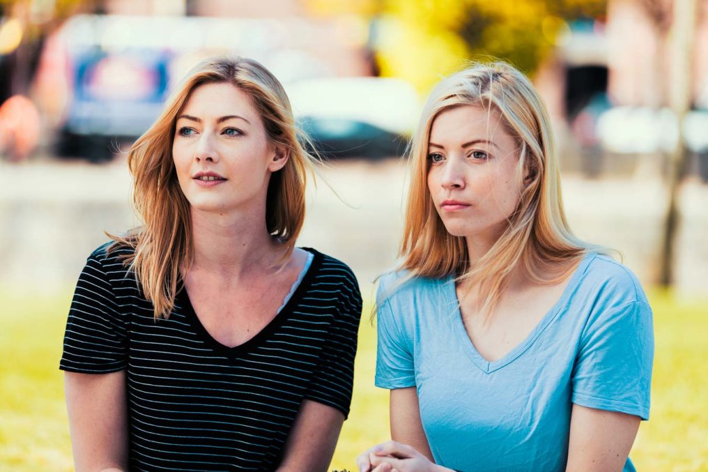 Two beautiful young women in casual clothes, with blond hair, look over to camera left with an out of focus urban background. The colours are of a cinematic quality