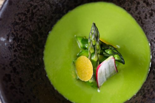 Green soup sits in a dark charcoal dappled bowl with asparagus tips and radish stacked in the centre