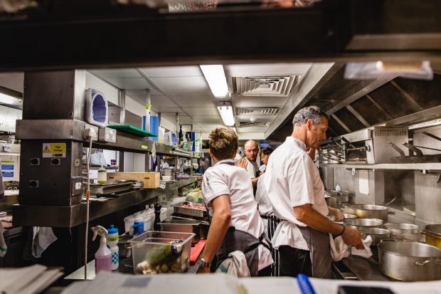 Chefs in the kitchen at Elystan Street. Two male chefs are reaching to each other while chef Phil Howard stands at the stove completely calm and unflustered