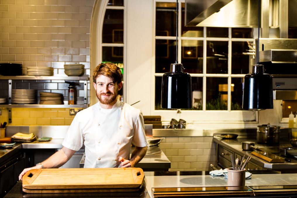 Environmental portrait of chef Lee Westcott at the pass of his kitchen at Typing Room, Bethnal Green, London. Young bearded male in chefs whites stands behind the serving pass of a fine dining restaurant. A night street scene can be seen through the window