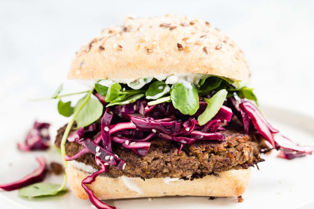 A front shot of a lentil veggie burger in a rustic seeded bun with shredded red cabbage, baby spinach and a white dressing