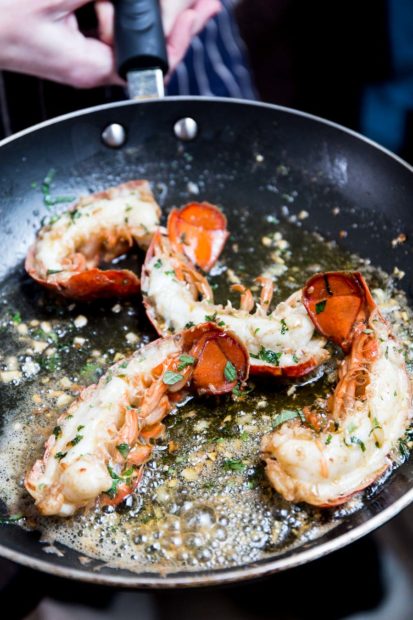 A close shot of someone holding a frying pan containing cooked lobster tails with bubbling garlic butter and green herbs