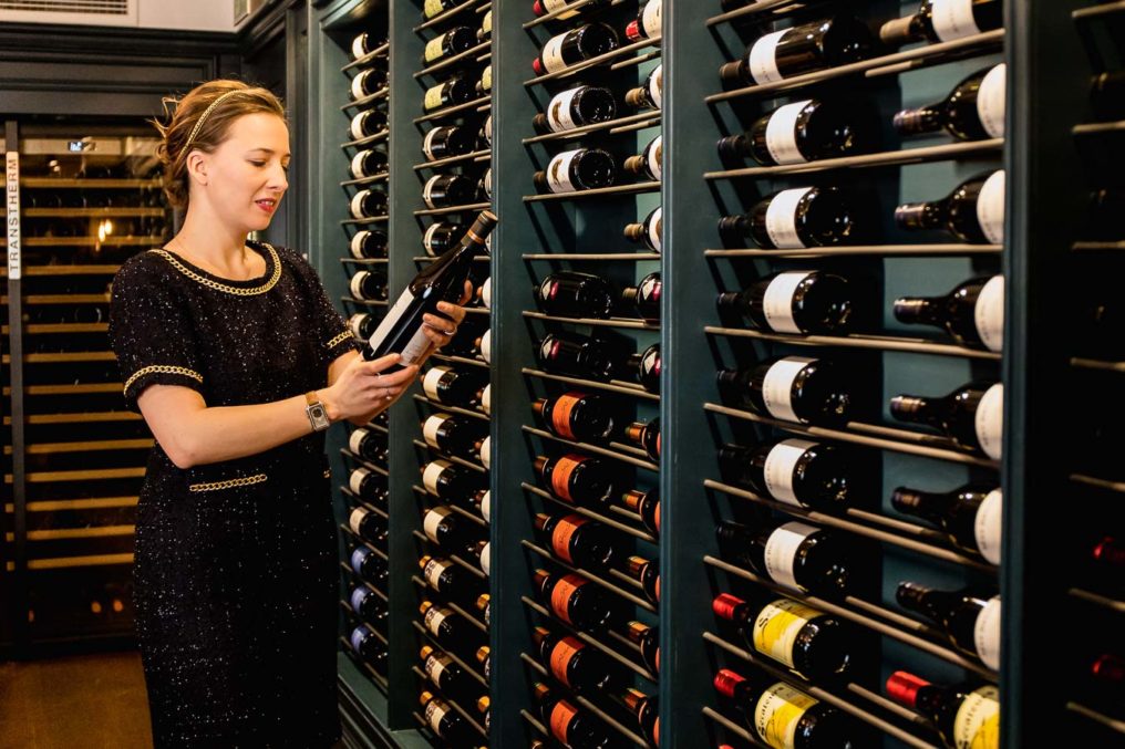 Lady in black and gold dress reading the wine label with a large wine rack along the whole wall in front of her. Margriet Crump, Sommelier