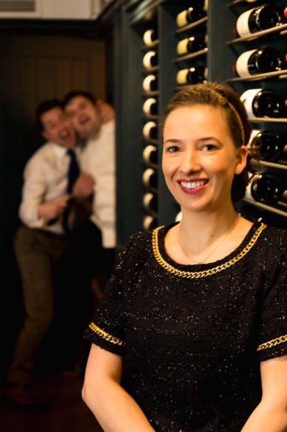 Portrait of Margriet Crump, Sommelier in the wine attic of The Oxford Blue Pub whilst Steven Ellis and Dan Crump joke around photo bombing in the background