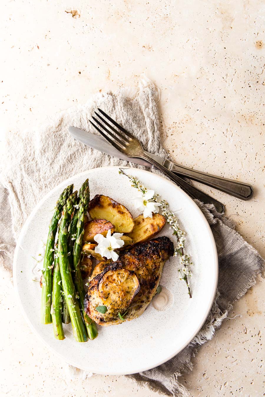 Flat lay of lemon roasted chicken on a white textured plate accompanied by roasted potatoes and asparagus, on a stone table top and textured cloth