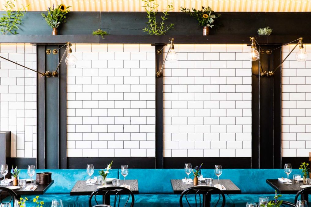 White tiled wall of a restaurant with dark ironwork pillars, blue brushed cotton seating, industrial style lights and potted plants. Paradise Garage by Robin Gill