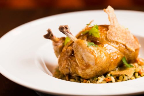 A golden, roasted poussin sitting on a bed of wild rice, all in a white, wide rimmed bowl