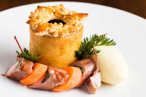 A pastry pie with an ornate pastry lid sits on a white plate surrounded by disc slices of meat and carrot alternating with a small rib meat and a large quenelle of mash potato