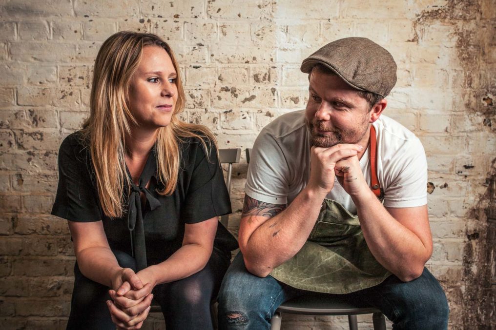 Fun portrait of Sarah and Robin Gill sitting side by side looking at each other with a rustic brick wall background