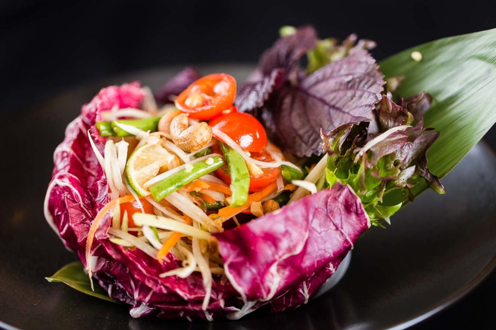 A dramatic photo of a Thai salad of cashews, tomato, green beans, sliced cabbage nestled in a large red cabbage leaf which sits on a black plate