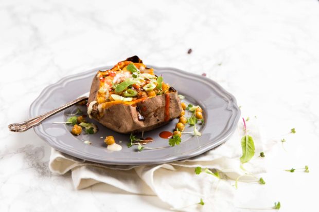 A sliced open baked sweet potato with a chick pea and spring onion tomato topping, on a grey plate with green micro herbs, a white linen textile and white marble table top