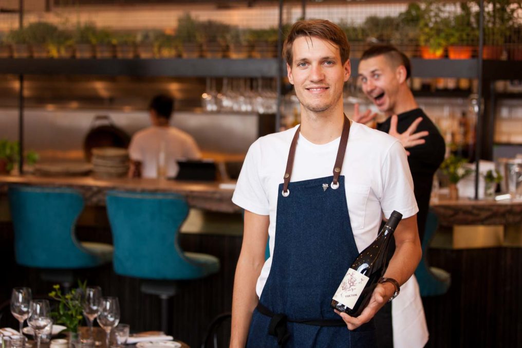 Environmental portrait of the Sommelier at Paradise Garage standing holding a bottle of wine while his colleague photobombs with a smile and jazz hands