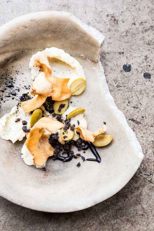 Dark coloured squid ink, prawn crackers and sliced olive sit on a partially broken, thick, stone bowl with restaurant styling and a stone floor tile as the background