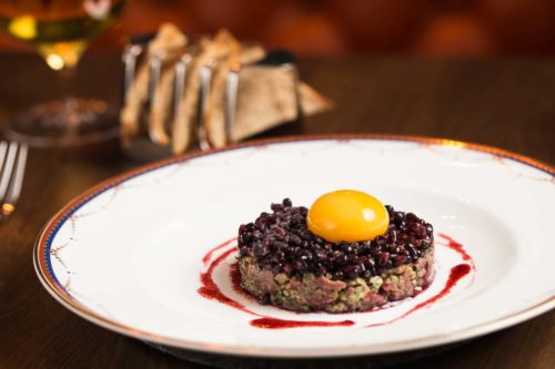 Gastro Pub style steak tartare, formed in a circle, topped with blackcurrants and the yoke of a hens egg on top. In the background is a wine glass and artisan toast crackers in a toast rack