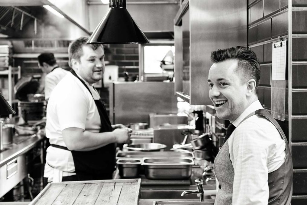 Candid fun photo in the kitchen of The Oxford Blue Pub, during service prep with Daniel Crump turning towards camera with a big smile and Chef Steven Ellis joining in from his prep station in the background