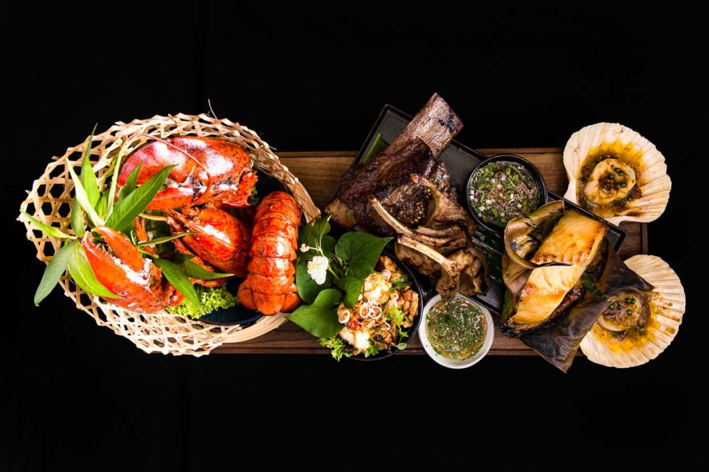 Top down, dramatic photo of a sharing seafood platter. Including a wicker basket of cooked lobster tails and claws, ribs, fish, dipping sauces and oysters in their shells. A celebration of surf and turf