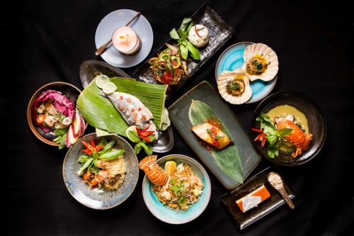 Top down shot of ten Thai dishes on separate plates in a black background. Scallops in shells, lobster tails, hake, King prawns, salads, white fish, desserts and lamb with rice