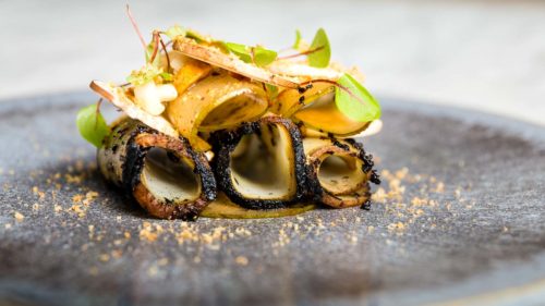 Fine dining from The Typing Room of potato rolled into tubes and blackened edges, laying on their sides, dressed with micro herbs and mushrooms on a grey textured plate