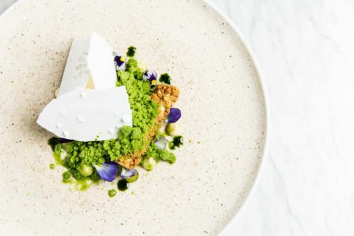 Fine dining plated dessert with white wafers hiding the ice-cream with a green crumb and edible flowers. The plate is white with speckles with a white marble background