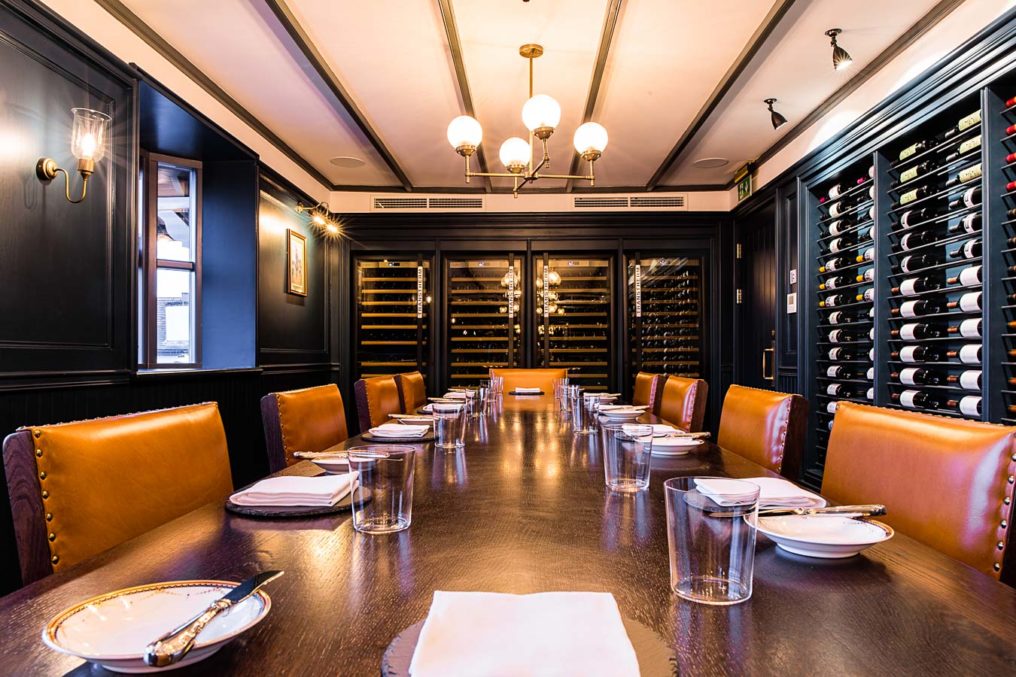 A long, wooden table stretches out lined with rich blown leather chairs, place setting all along the table. The open wine rack lines the right hand wall of the wine attic at The Oxford Blue Pub and four wine fridges can be seen at the far end of the room which is lit be wall and ceiling lights