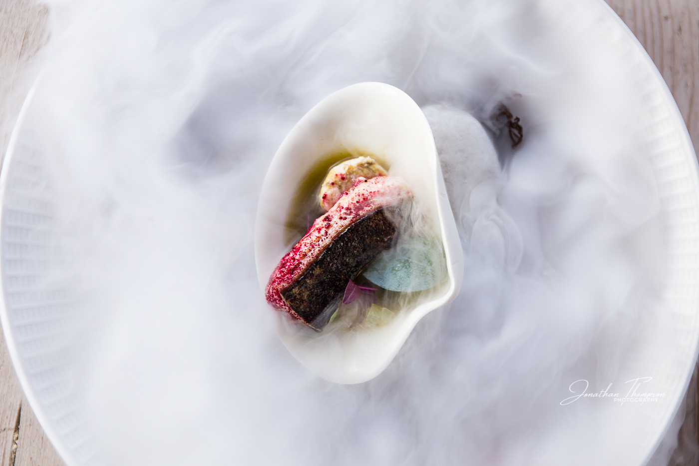 Top down view of a piece of cooked monk fish in a white bowl which the smoke from dry ice surrounding it. Chester Zoo