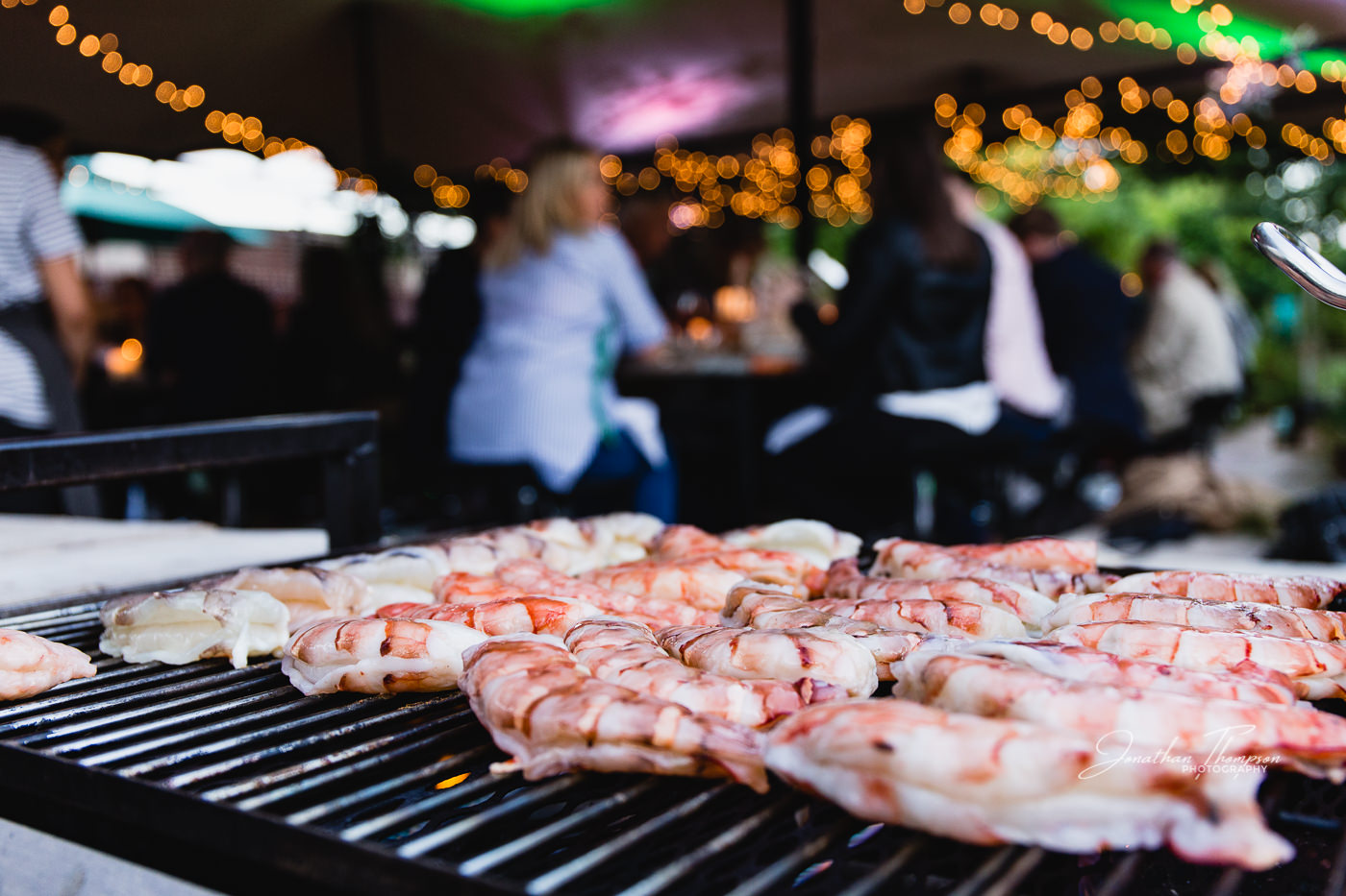 Low angle view of king prawns laying side by side on a bbq grill with diners sitting at tables in the background. Chester Zoo
