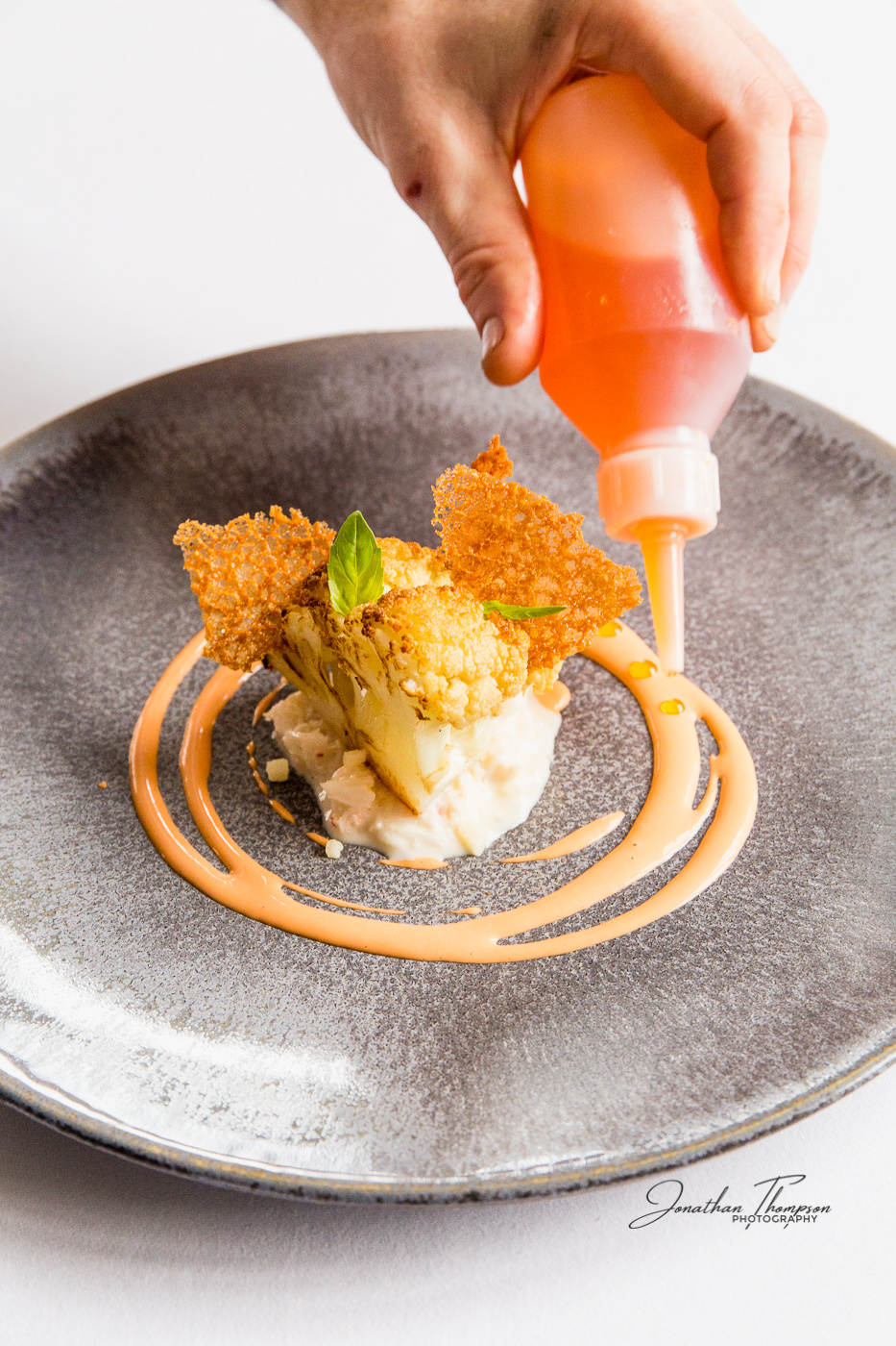 A squeezy bottle dropping small amounts of oil into a sauce. The plate is grey containing a cauliflower dish. Food design by chef Lee Westcott
